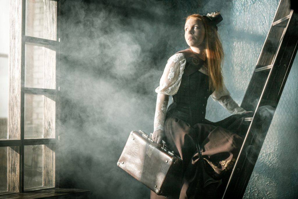 Redhead young woman dressed in steampunk style is standing one foot on a wooden ladder in cloud of steam. She holding a old suitcase in her hand and looking over shoulder on a large window