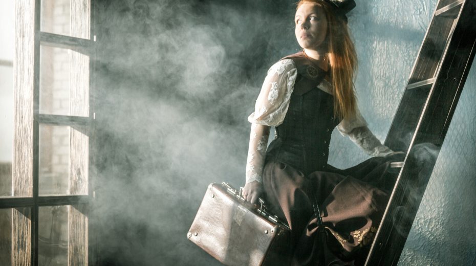 Redhead young woman dressed in steampunk style is standing one foot on a wooden ladder in cloud of steam. She holding a old suitcase in her hand and looking over shoulder on a large window