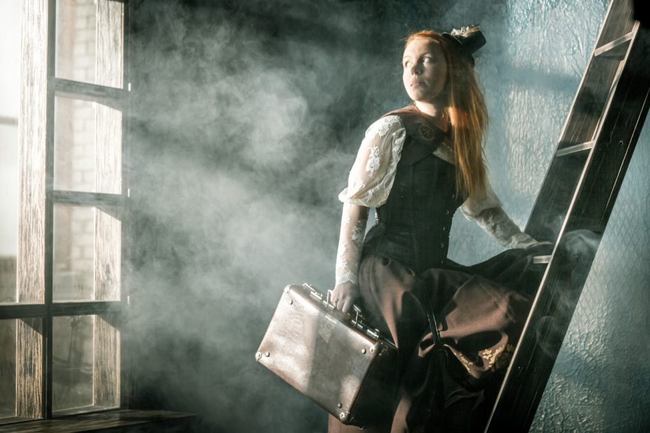 Redhead young woman dressed in steampunk style is standing one foot on a wooden ladder in cloud of steam. She holding a old suitcase in her hand and looking over shoulder on a large window